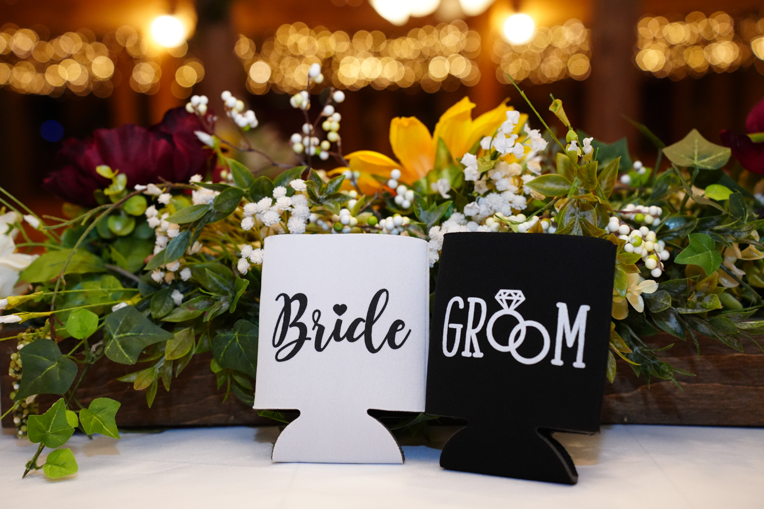 Bride and groom koozies sitting on the head table of a reception area with warm string lights behind them