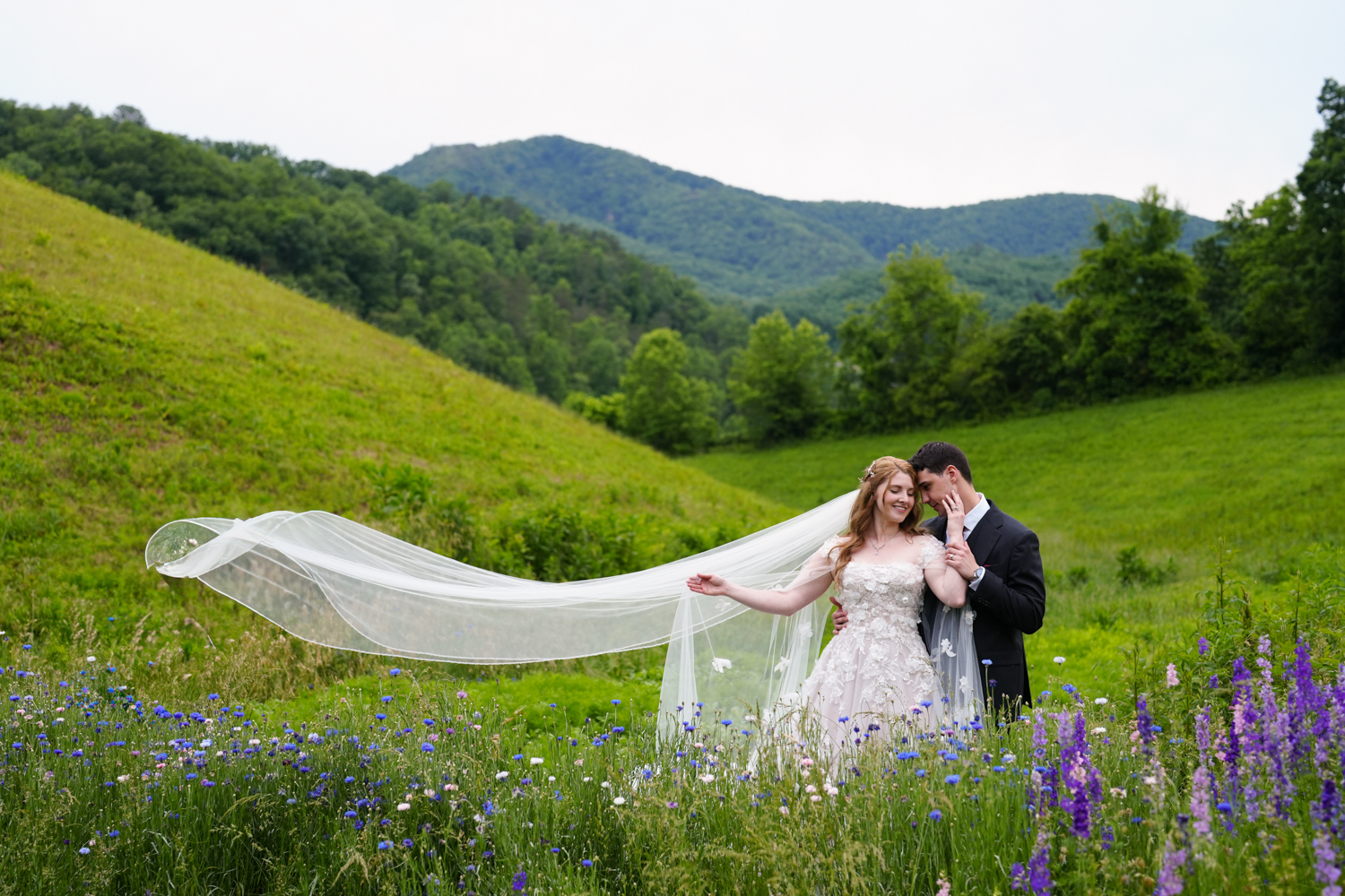 Wedding portrait with a bride's veil blowing in the wind as she caresses her groom's face in a wildflower garden with a mountain ridge view