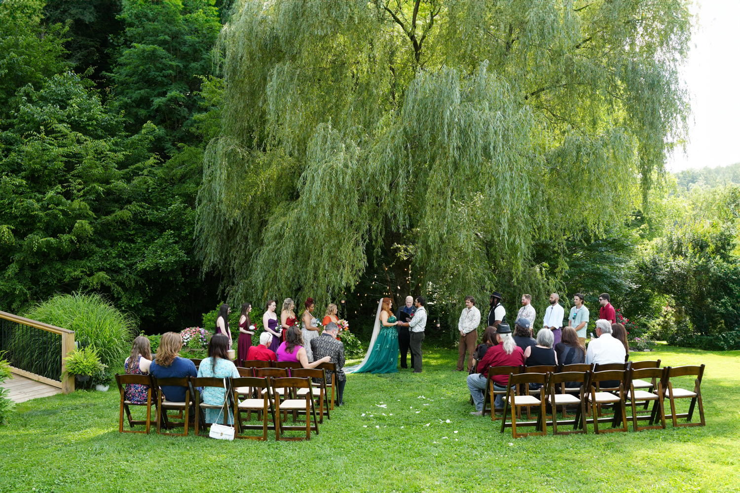 Wedding ceremony with a forest theme under the willow tree at Honeysuckle Hills with a bride in a green wedding dress and guests seated in wooden folding chairs