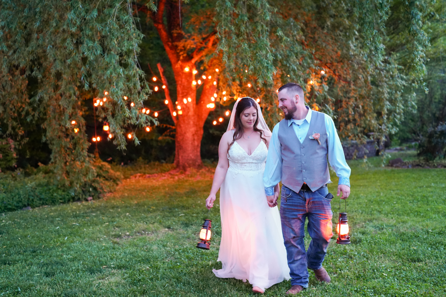 Wedding couple at dusk carrying lanterns in front of a willow tree with golden string lights in the evening at Honeysuckle Hills